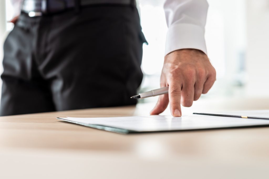 Businessman standing next to his office desk holding a pen pointing on a document or subscription form to where the signature line is.